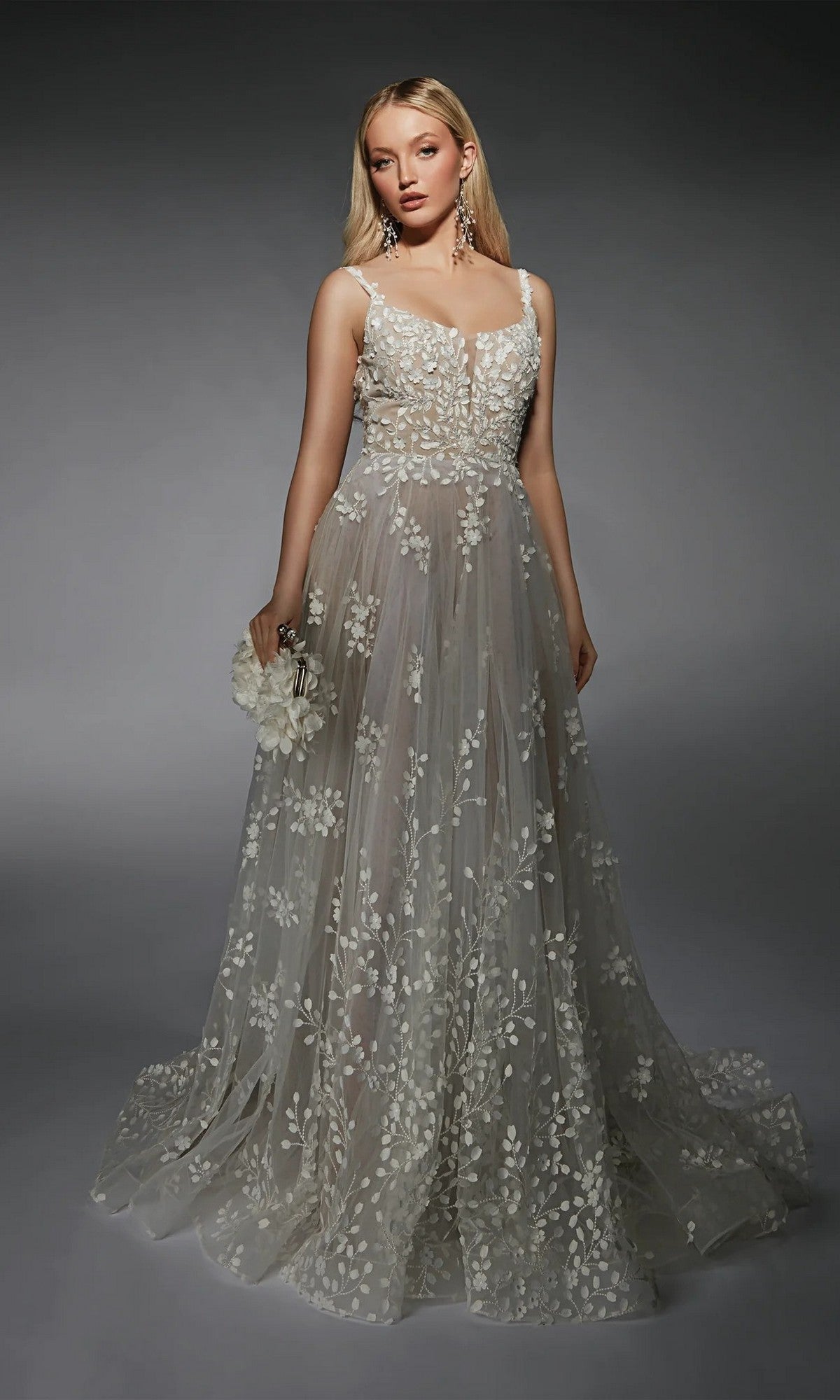 A woman wears the Long White Dress: Alyce 7132 in Ivory/Lt Latte—a sleeveless, floor-length gown with sheer fabric and delicate white floral embroidery—holding a small flower accessory against a dark backdrop.